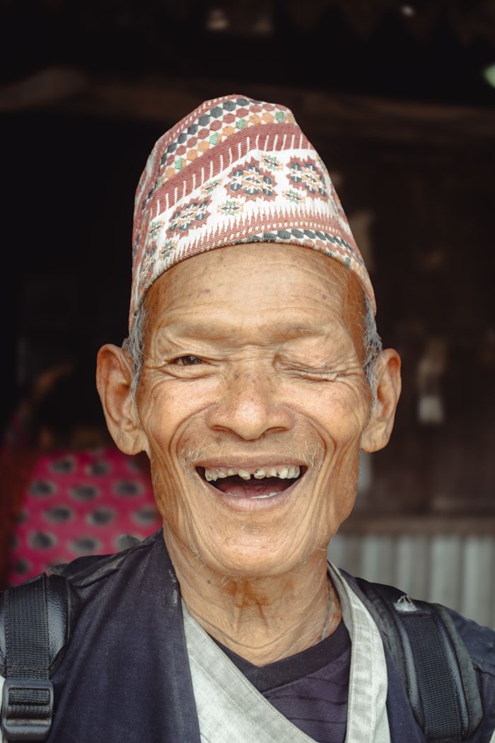 Portrait of an elderly man joyfully smiling, wearing a patterned traditional hat, with a rustic background.